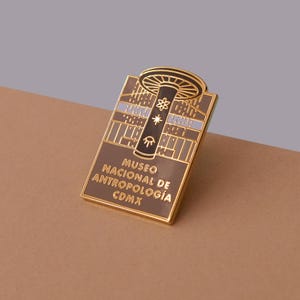 May include: A gold-colored enamel pin featuring a brown and white illustration of the Museo Nacional de Antropología in Mexico City. The pin has the text "Museo Nacional de Antropología CDMX" on it.
