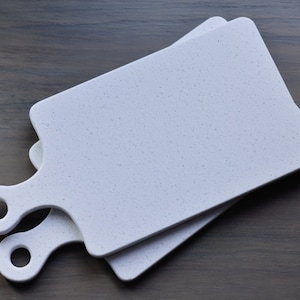 May include: Two white cutting boards with speckled surfaces and rounded handles. The boards are stacked on top of each other.
