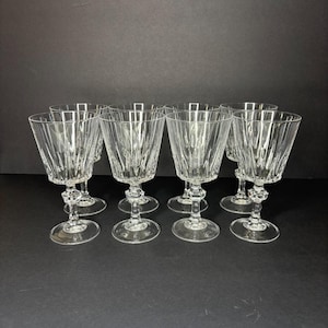 May include: Set of eight clear crystal wine glasses. Each glass features a faceted bowl design, a decorative stem, and a round base. The glasses are arranged on a dark surface, showcasing their elegant design.