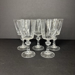 May include: A set of six clear crystal wine glasses. Each glass features a faceted bowl, a decorative stem, and a round base. The glasses are arranged on a dark surface, showcasing their elegant design and clarity.