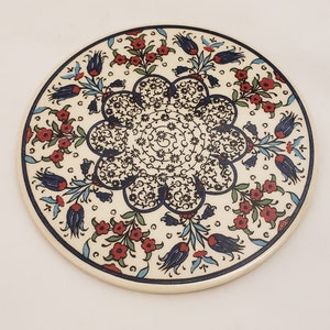 May include: A round ceramic plate with a white background and a blue and red floral design. The design features a central motif of a stylized flower with intricate details, surrounded by smaller flowers and leaves.