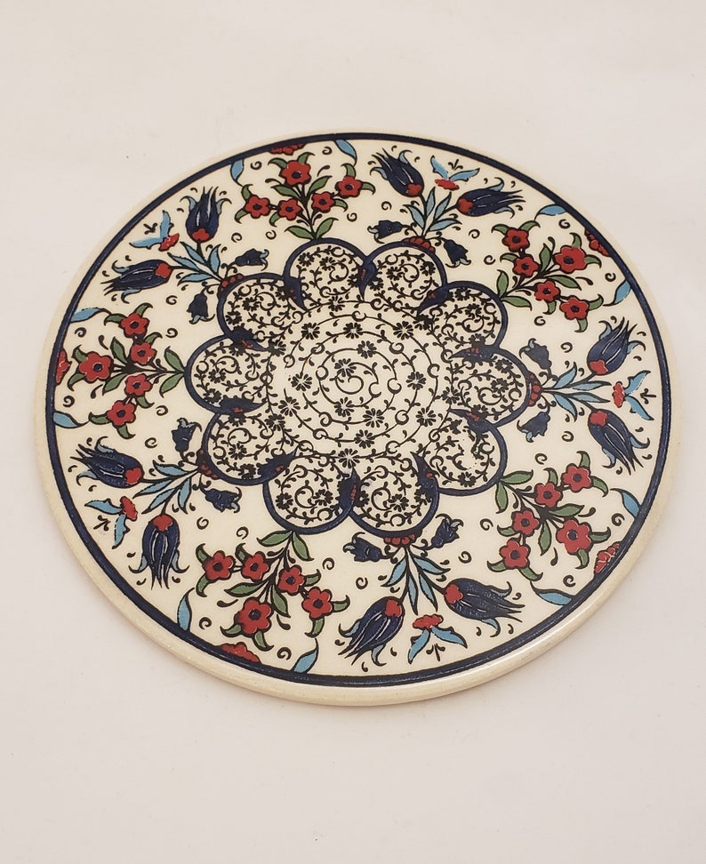 May include: A round ceramic plate with a white background and a blue and red floral design. The design features a central motif of a stylized flower with intricate details, surrounded by smaller flowers and leaves.