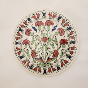May include: A white ceramic plate with a red, green, and blue floral design. The design features stylized carnations and tulips.