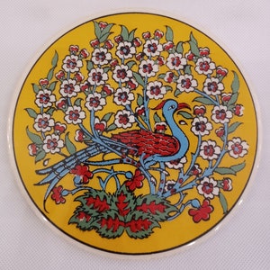Puede incluir: Un azulejo de cerámica redondo con fondo amarillo y un pájaro azul y rojo posado en una rama con flores blancas. El pájaro está rodeado de hojas verdes y flores rojas.