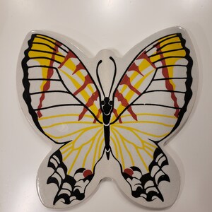 May include: A white ceramic butterfly wall decor with black, yellow, and red painted details. The butterfly has a stylized design with a black outline and yellow and red stripes on the wings.
