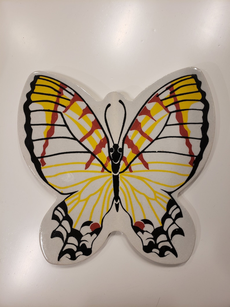 May include: A white ceramic butterfly wall decor with black, yellow, and red painted details. The butterfly has a stylized design with a black outline and yellow and red stripes on the wings.
