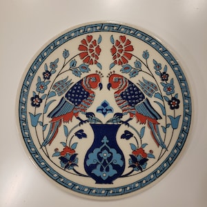 May include: A round ceramic plate with a white background and blue and red floral designs. Two red birds with blue and white patterns are perched on a blue and white vase.