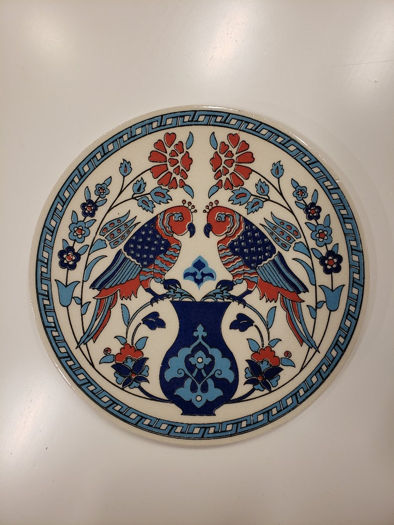 May include: A round ceramic plate with a white background and blue and red floral designs. Two red birds with blue and white patterns are perched on a blue and white vase.