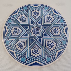 May include: A round ceramic coaster with a blue and white geometric pattern. The pattern features stars, diamonds, and other shapes.