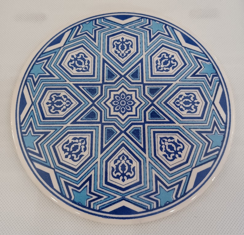 May include: A round ceramic coaster with a blue and white geometric pattern. The pattern features stars, diamonds, and other shapes.