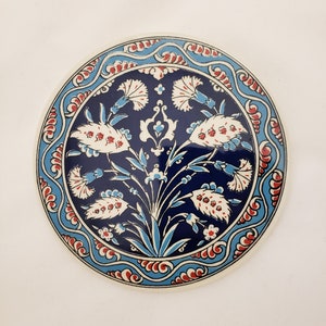 May include: A round ceramic coaster with a blue and white floral design. The design features stylized flowers with red accents and a blue background. The coaster has a white border.