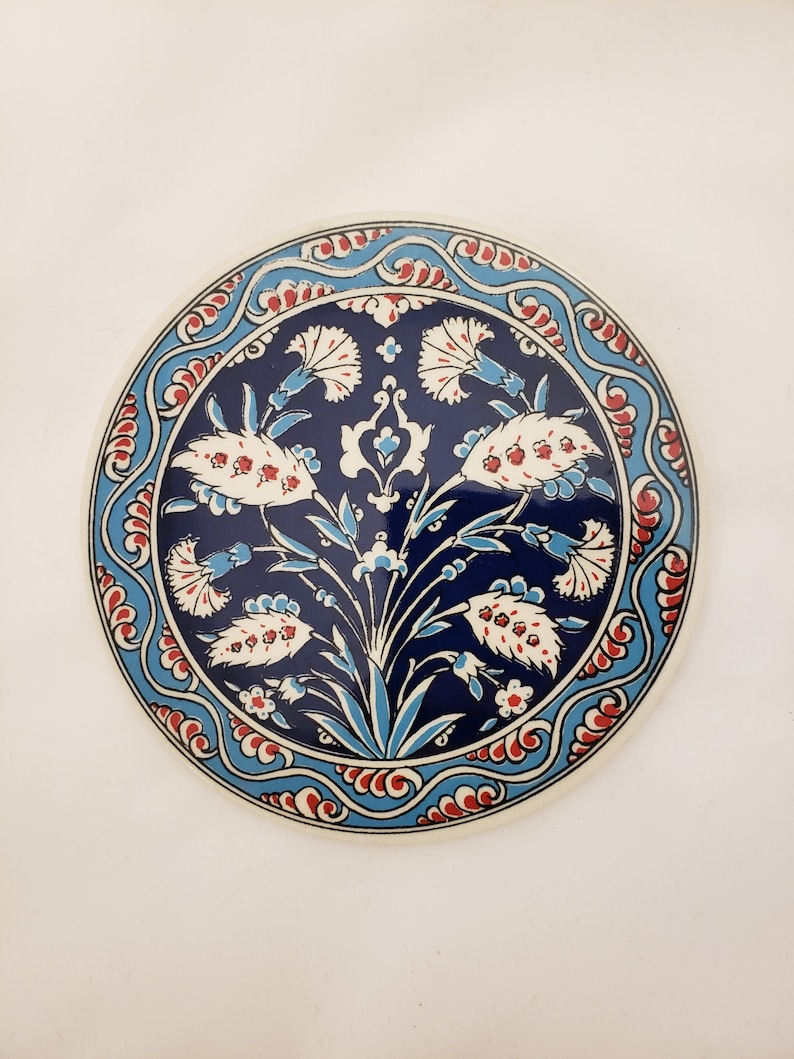 May include: A round ceramic coaster with a blue and white floral design. The design features stylized flowers with red accents and a blue background. The coaster has a white border.