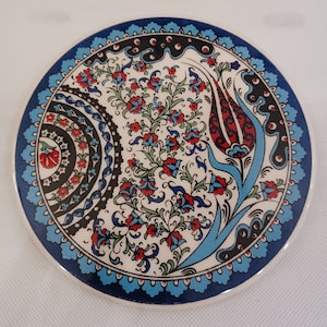 May include: A round ceramic coaster with a blue and white border and a floral design in the center. The design features red, blue, and green flowers and leaves.