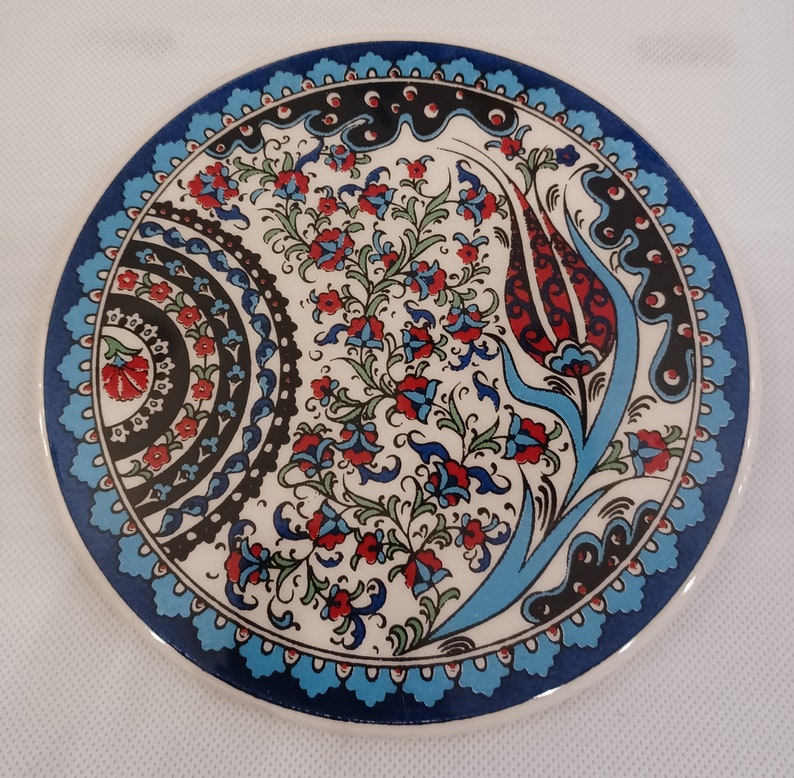 May include: A round ceramic coaster with a blue and white border and a floral design in the center. The design features red, blue, and green flowers and leaves.
