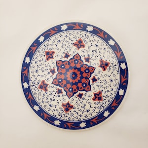 May include: A round ceramic coaster with a white background and a blue and red floral design. The design features a central starburst with intricate details.