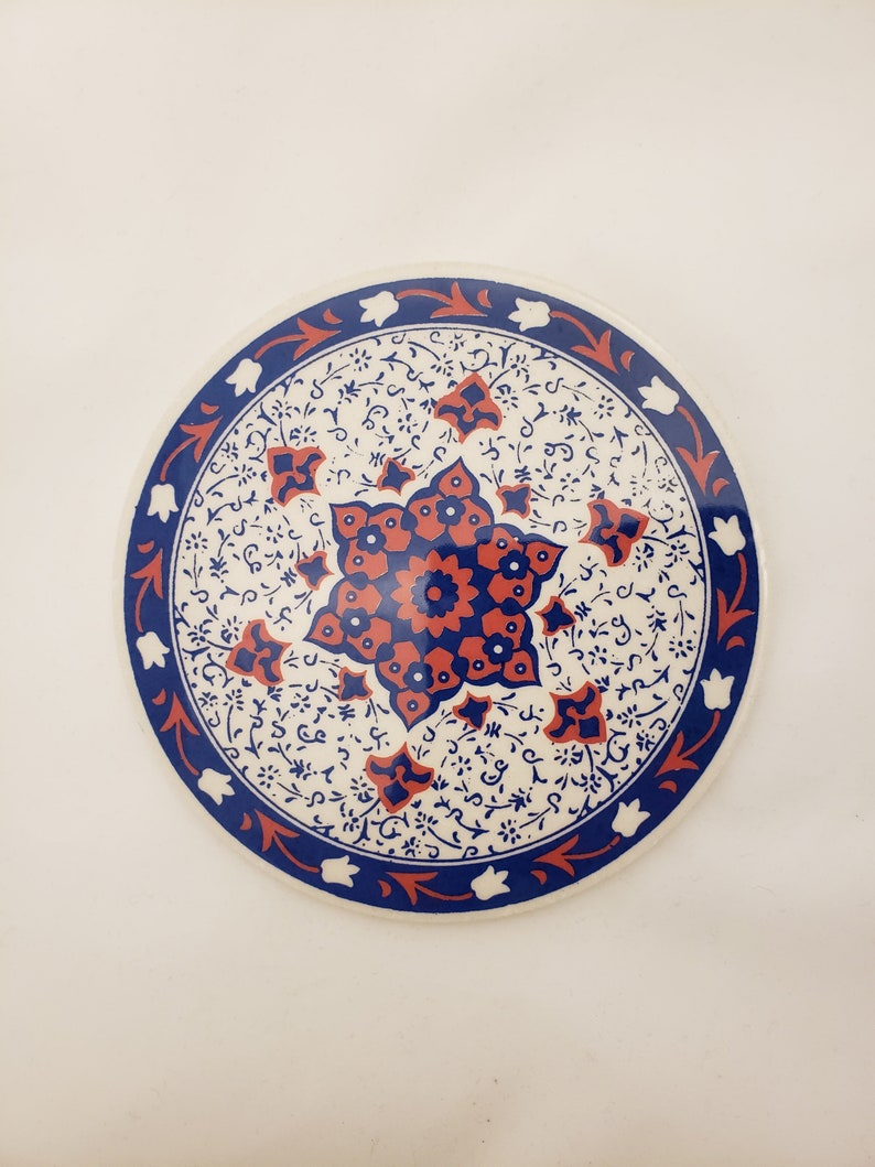 May include: A round ceramic coaster with a white background and a blue and red floral design. The design features a central starburst with intricate details.