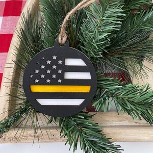 May include: Black wooden Christmas ornament shaped like a circle with a thin yellow stripe across the middle and a white and black American flag design above the stripe.