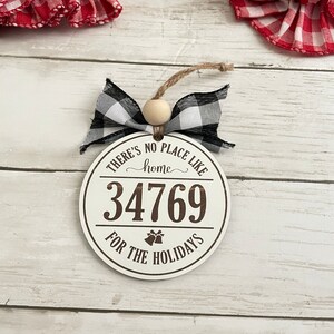 May include: White round wooden ornament with the text "There's no place like home for the holidays" and the number "34769". The ornament is decorated with a black and white buffalo plaid bow.