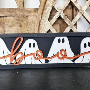 May include: A black framed wooden sign with four white ghosts holding an orange ribbon that spells out "boo".