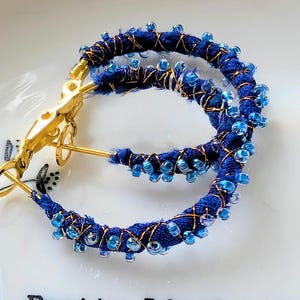 May include: A pair of blue hoop earrings. The hoops are wrapped in blue fabric and decorated with small blue beads. Gold-coloured metal accents the design. The earrings are on a white surface with the text "Pretty little things".