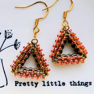 May include: Handmade beaded triangle earrings in shades of brown, orange, and gold. The earrings are suspended from gold-coloured ear wires. The text "Pretty little things" is printed below the earrings. The earrings are a unique accessory.
