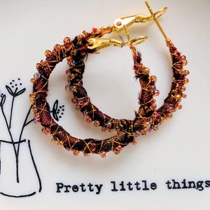 May include: A pair of hoop earrings with a dark brown fabric wrapped around the hoops, accented with gold wire and small, iridescent beads. The earrings have gold-coloured clasps. The earrings are on a white plate with the text "Pretty little things".