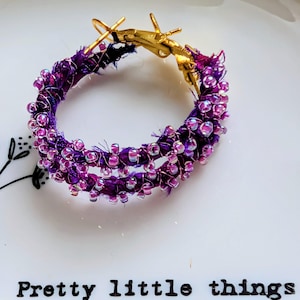 May include: A single hoop earring with a gold clasp and a purple beaded design. The earring is adorned with small, iridescent beads and wrapped in a dark purple fabric. The text "Pretty little things" is printed on the surface.