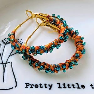 May include: Gold hoop earrings with orange fabric and teal bead detailing. The earrings are presented on a white plate with the words "Pretty little things".