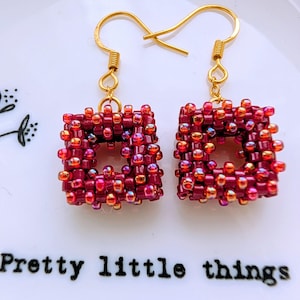 May include: A pair of handmade square earrings. The earrings are made of small, red and gold beads, with a gold-colored hook. The earrings are on a white surface with the text "Pretty little things".
