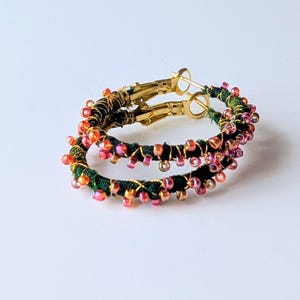 May include: A pair of hoop earrings. The hoops are wrapped in dark green fabric and decorated with small pink, orange, and gold beads. The earring clasps are gold-coloured.
