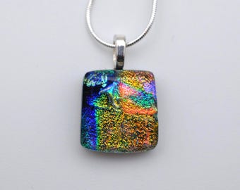 May include: A square dichroic glass pendant necklace. The pendant is a mosaic of colors including blue, green, yellow, and orange. The pendant is attached to a silver chain. The pendant is approximately 0.75 inches square.