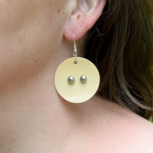 Puede incluir: Un par de pendientes redondos de color crema con detalles plateados. Los pendientes cuelgan de la oreja de una persona.