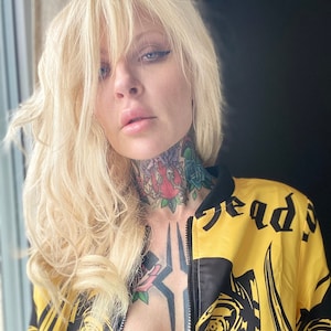 May include: A woman with blonde hair is wearing a yellow and black bomber jacket with a skull graphic and the word "Heads" printed on the back. The jacket is unzipped, revealing a black bra and tattoos on her chest and neck.