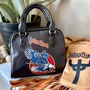 May include: Black handbag with a Judas Priest logo and an eagle graphic. The bag has a gold handle and a brown fringe.