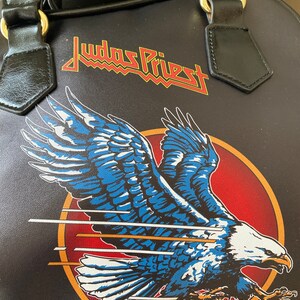 May include: Black leather handbag with a red and yellow "Judas Priest" logo and a blue and white eagle graphic.