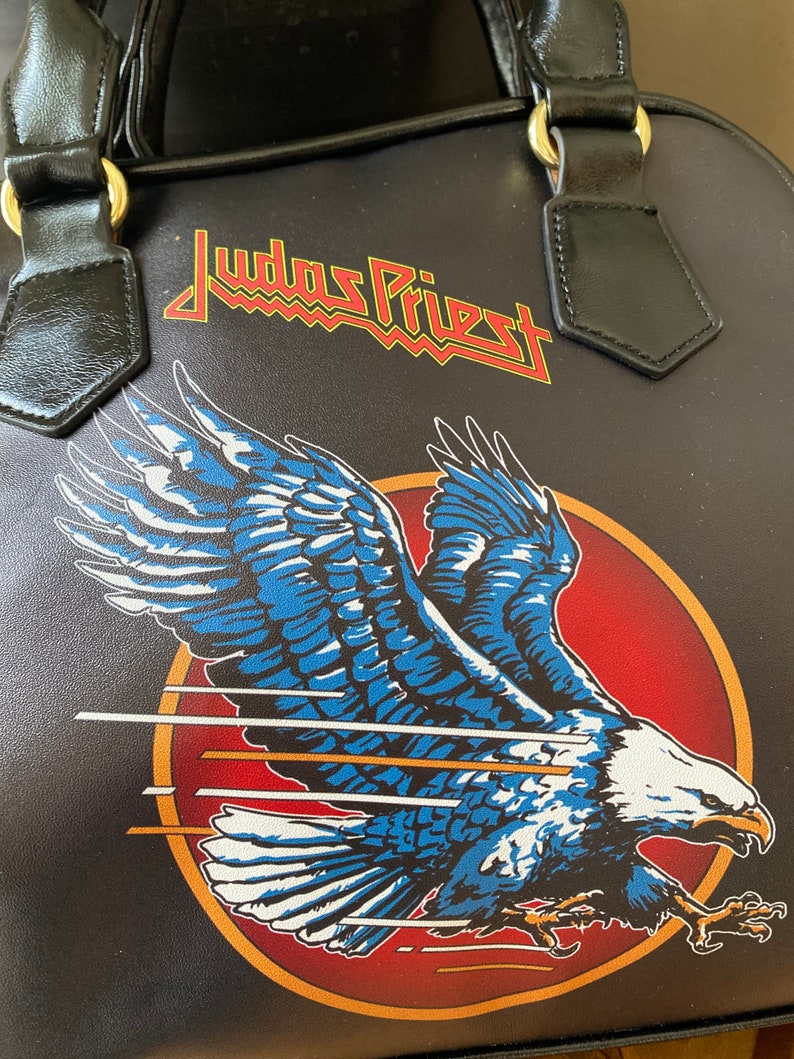 May include: Black leather handbag with a red and yellow "Judas Priest" logo and a blue and white eagle graphic.