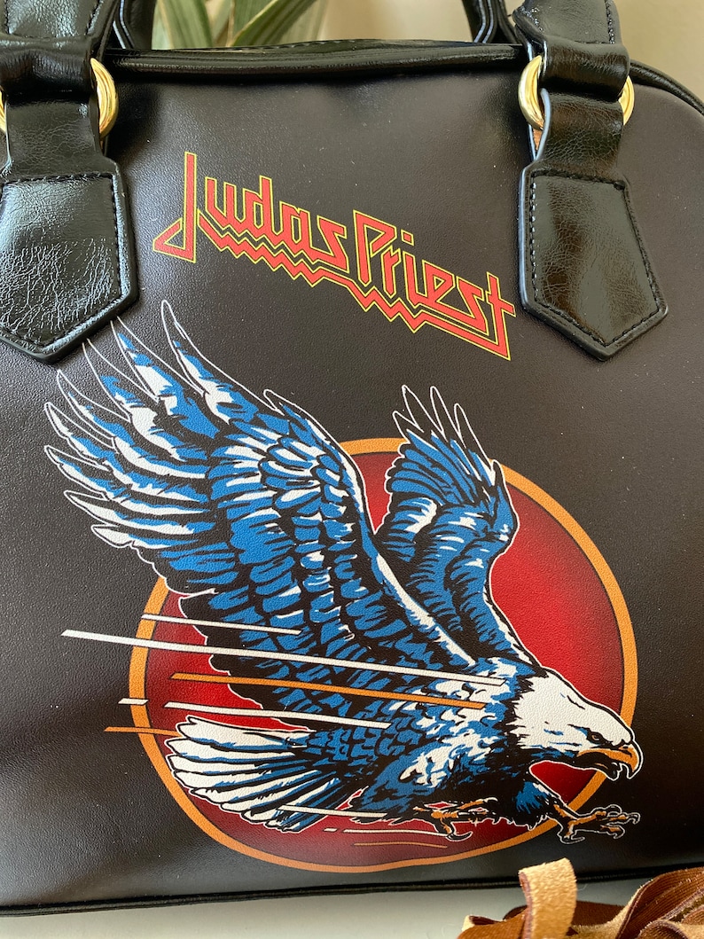 May include: Black leather handbag with a Judas Priest logo and a flying eagle graphic. The eagle is blue, white, and red.