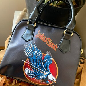 May include: Black leather handbag with a Judas Priest logo and a flying eagle design. The eagle is blue, white, and red.