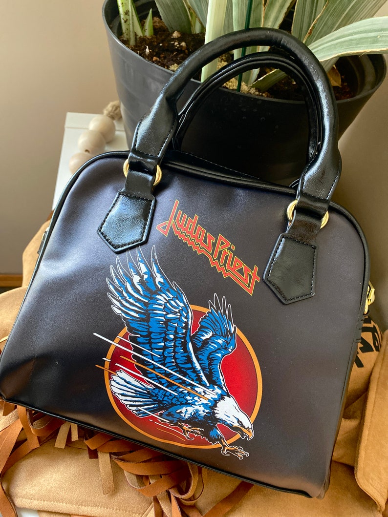 May include: Black leather handbag with a Judas Priest logo and a flying eagle design. The eagle is blue, white, and red.