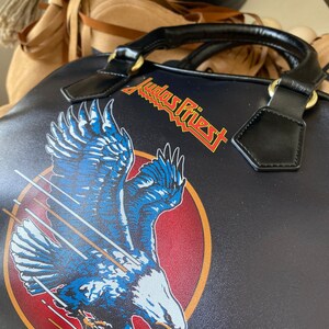 May include: Black leather handbag with a red, white, and blue eagle design and the text "Judas Priest" in yellow lettering.