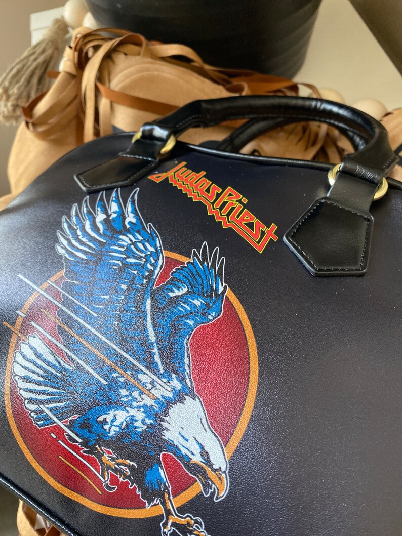 May include: Black leather handbag with a red, white, and blue eagle design and the text "Judas Priest" in yellow lettering.