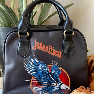 May include: Black leather handbag with a Judas Priest logo and an eagle design. The bag has a top handle and a detachable shoulder strap.