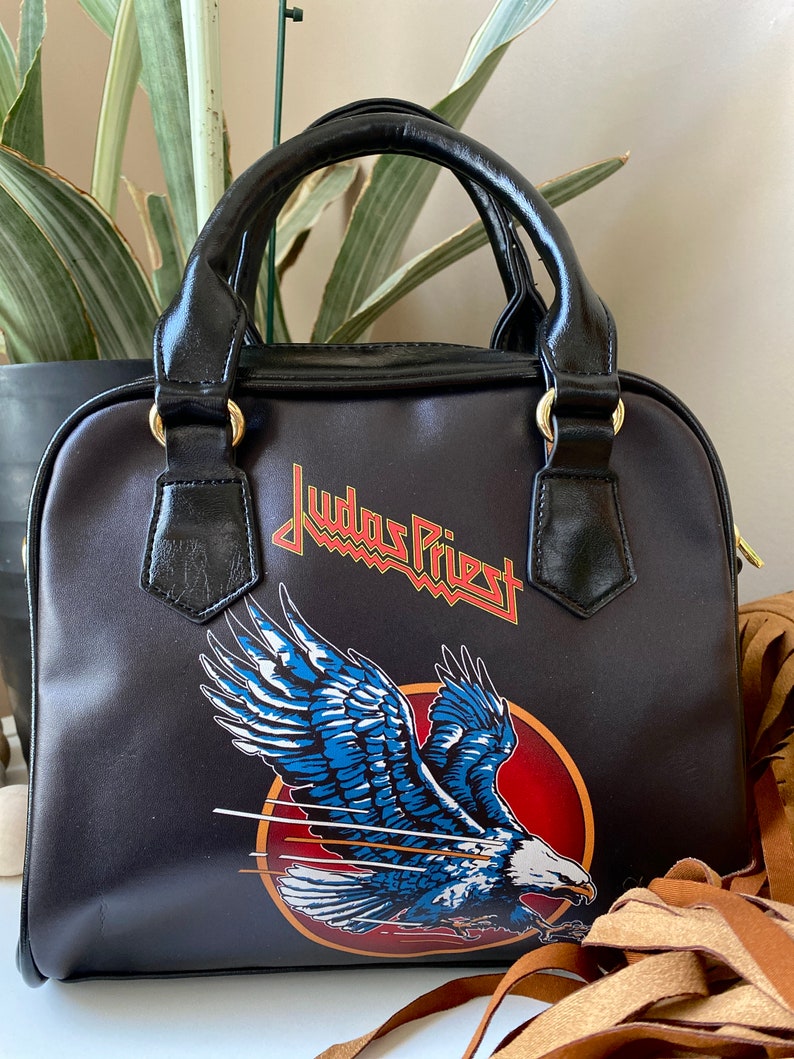 May include: Black leather handbag with a Judas Priest logo and an eagle design. The bag has a top handle and a detachable shoulder strap.