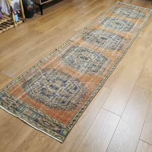 May include: A long, rectangular rug with a faded orange background and intricate blue and green patterns. The rug has a fringed edge and is laid out on a wooden floor.