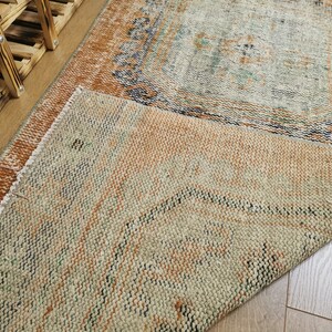 May include: A close-up of a folded rug with a beige, brown, and green pattern. The rug has a distressed look and is made of a woven material.
