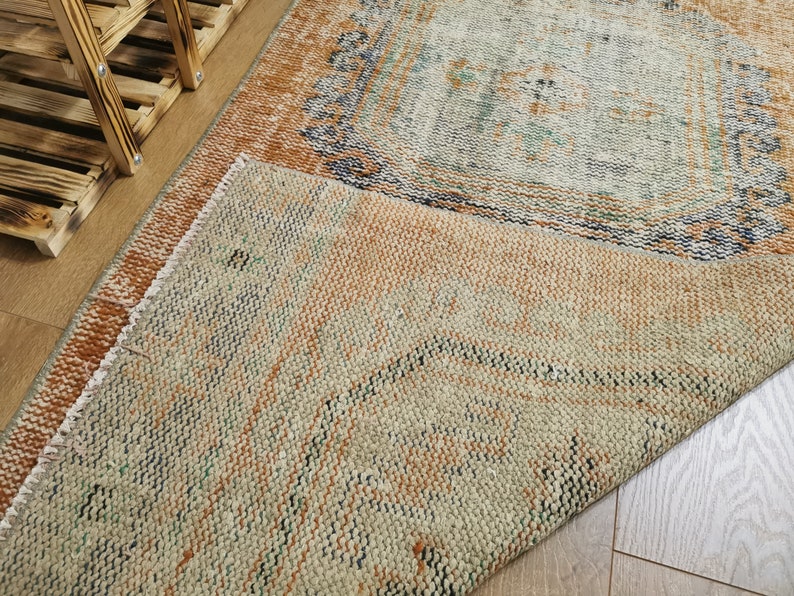 May include: A close-up of a folded rug with a beige, brown, and green pattern. The rug has a distressed look and is made of a woven material.