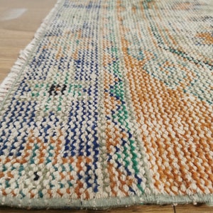 May include: Close-up of a woven rug with a fringe edge. The rug has a cream background with stripes of orange, blue, and green.