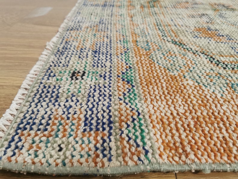 May include: Close-up of a woven rug with a fringe edge. The rug has a cream background with stripes of orange, blue, and green.