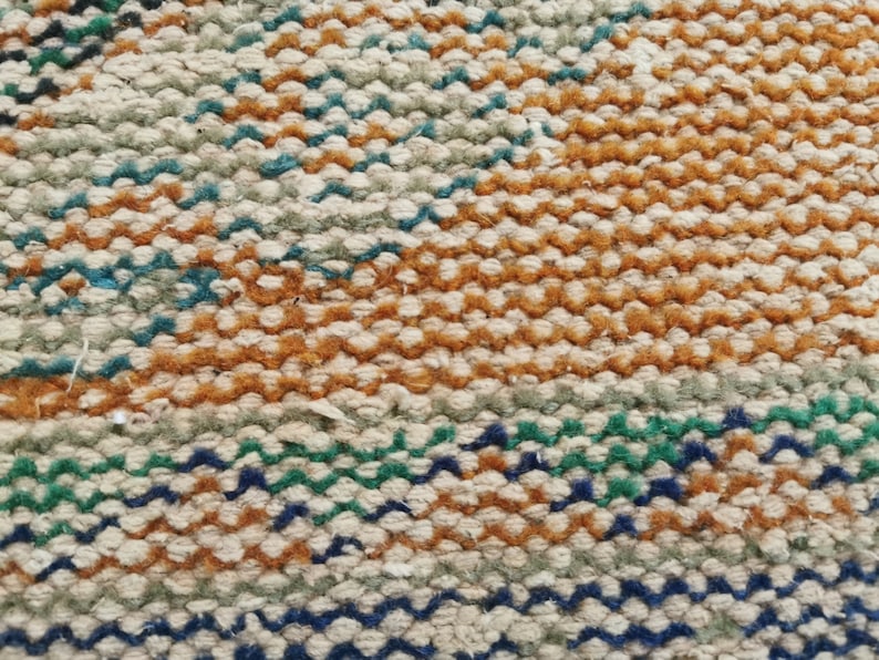May include: Close-up of a woven rug with a multi-colored pattern. The rug is made of natural fibers and has a textured surface. The colors in the pattern are brown, beige, green, and blue.