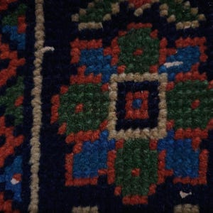 Op de afbeelding: Close-up van een deel van een donkerblauw tapijt met een herhalend bloemmotief. Het patroon heeft groene, rode en blauwe bloemmotieven met een beige rand. Het tapijt heeft een getextureerd uiterlijk.